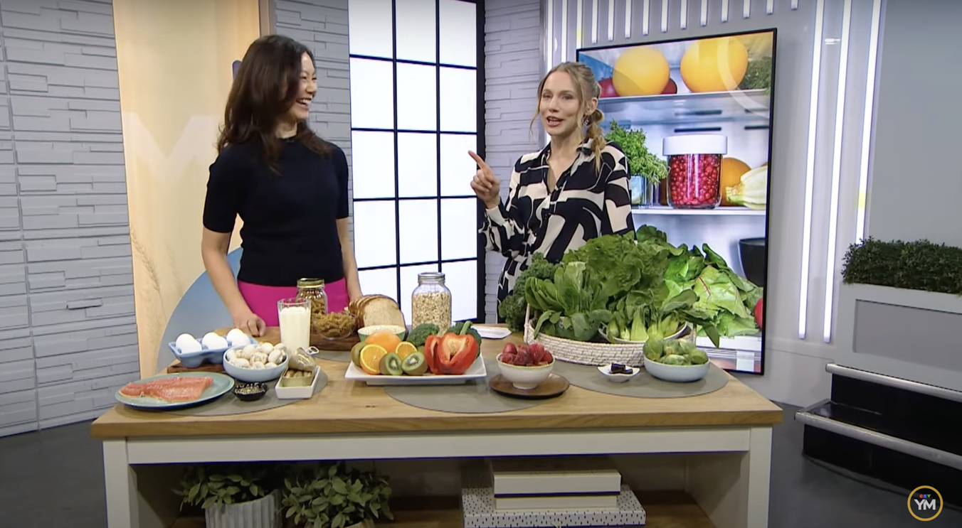 Sue is standing in the TV studio with TV host Kelsey McEwan. On the table are foods such as milk, salmon, eggs, mushrooms, vitamin D supplements; whole grain breads and pasta; strawberries, broccoli, red pepper, orange, kiwis; basket of leafy green vegetables; small dish of dark chocolate