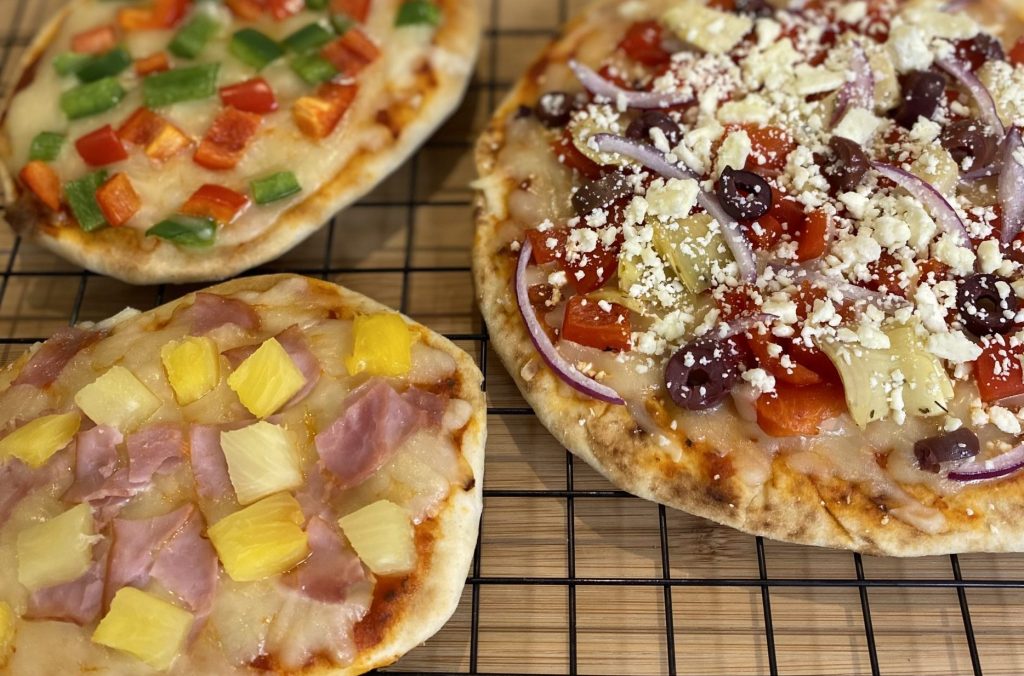 3 pizzas made on naan. One pizza has red and green peppers; one pizza has ham and pineapple; one pizza has olives, roasted red peppers, artichokes, red onion and feta cheese.