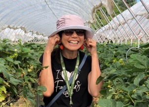 Sue smiling in a strawberry field and holding 2 strawberries as earrings
