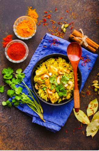 A plate of Chicken Biryani on a bright blue napkin, surrounded by fresh herbs, spices and a wooden spoon.