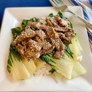 A plate of rice, topped with baby bok choy, beef slices and sesame seeds.