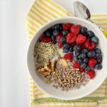 A bowl of cooked oatmeal topped with walnuts, hemp seeds, bran cereal, blueberries and raspberries. The bowl is on a yellow towel with a yellow spoon.