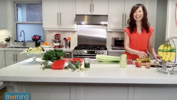 Sue wearing an orange dress and standing behind her kitchen counter which if filled with fruit and veggie props.