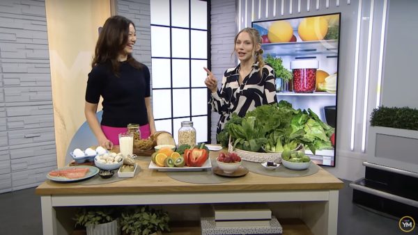 Sue is standing in the TV studio with TV host Kelsey McEwan. On the table are foods such as milk, salmon, eggs, mushrooms, vitamin D supplements; whole grain breads and pasta; strawberries, broccoli, red pepper, orange, kiwis; basket of leafy green vegetables; small dish of dark chocolate