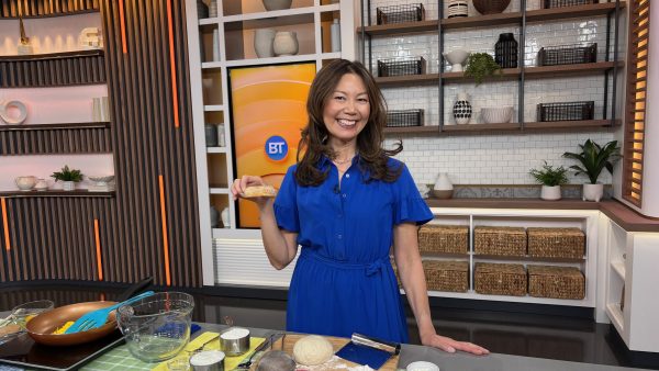 Sue is standing behind a TV set counter and holding half a home made bagel
