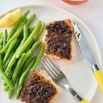 A white plate with a piece of salmon cut in half, green beans, wedge of lemon and yellow cutlery.